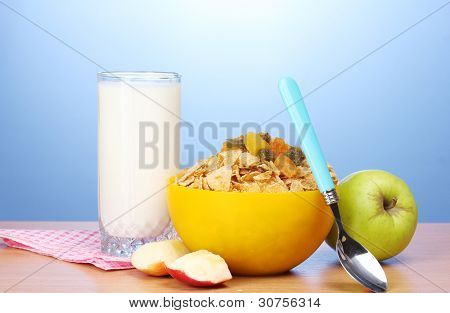leckere Cornflakes in gelbe Schale, Äpfel und Glas Milch auf Holztisch auf blauem Hintergrund