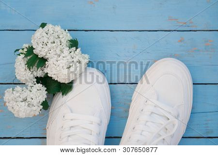 The Toes Of White Women’s Sneakers Lie On A Blue Wooden Background Next To A Bouquet Of White Flower