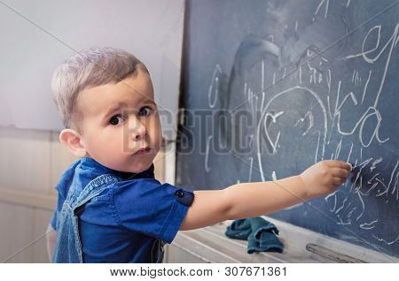 Little Boy Standing Near School Blackboard With A Piece Of Chalk