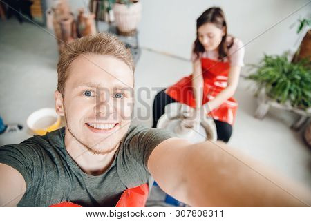 Happy Man Makes Selfie Photo On Background Of Girl Behind A Potter Wheel, Dishes From Clay. Concept 