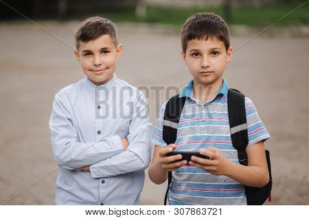 Two Boy Sitting On The Bench And Play Online Games. One Boys With Backpack. Young Boys Use Their Pho