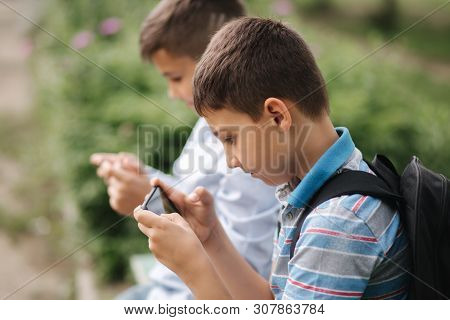 Two Boy Sitting On The Bench And Play Online Games. One Boys With Backpack. Young Boys Use Their Pho