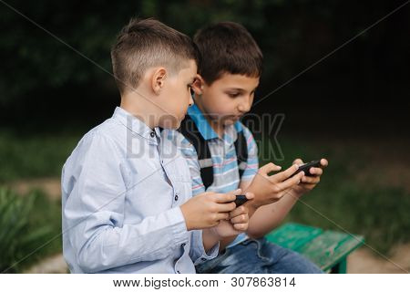 Two Boy Sitting On The Bench And Play Online Games. One Boys With Backpack. Young Boys Use Their Pho