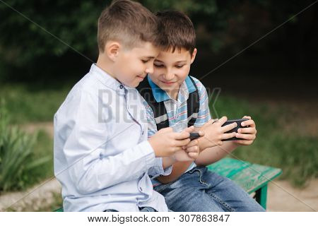 Two Boy Sitting On The Bench And Play Online Games. One Boys With Backpack. Young Boys Use Their Pho