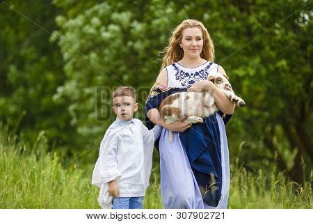 Portrait Of Smiling Mother With Her Little Son. Holding Cub Of Newborn Lamb Outdoors On Nature Backg