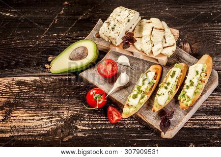 Bruschetta With Grilled Cheese Halumi, Avocado And Cherry On A Wooden Background. Preparation Of Che