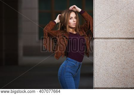 Young fashion woman walking on city street Stylish female model in brown leather fringe suede jacket and dark blue jeans