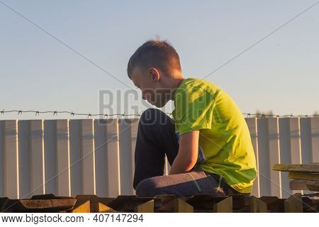 A Sad Boy Sits On The Roof Of A Barn Near A Fence With Barbed Wire In The Summer