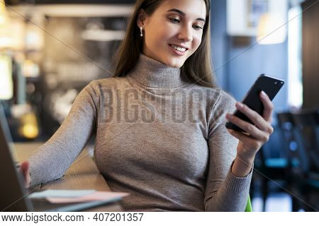 Attractive Business Woman Sits At Table In Front Of Laptop And Talks On Mobile Phone, Negotiates On 