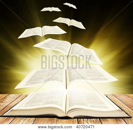 Offenes Buch auf hölzernen Planken und Seiten fliegen bis auf schwarzem Hintergrund mit Brigth Strahlen