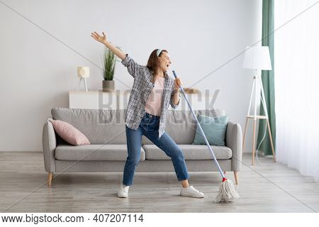 Happy Young Woman Singing While Washing Floor At Living Room, Using Mop As Microphone, Having Fun Du