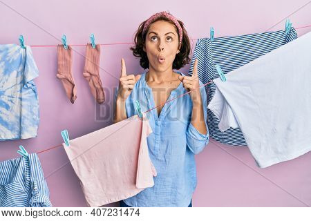 Young brunette woman doing laundry around string hangs amazed and surprised looking up and pointing with fingers and raised arms. 