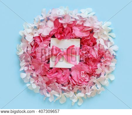 White Gift Box With A Bow Between Pink Flowers Top View On Light Blue Background
