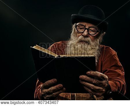 A wise old man with a long gray beard in a bowler hat and glasses is reading an old book. Black background. 