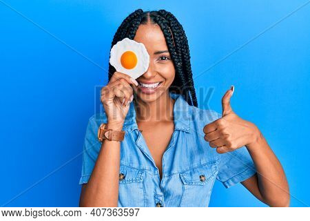 Beautiful hispanic woman holding fried egg smiling happy and positive, thumb up doing excellent and approval sign 