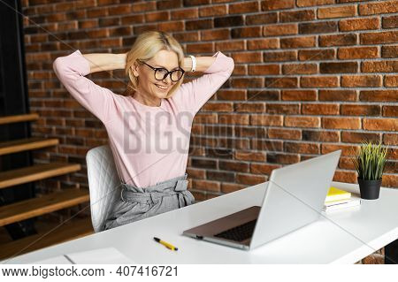 Mature Mid-age Businesswoman Sitting At The Desk, Looking At The Laptop And Smiling, Excited, Satisf