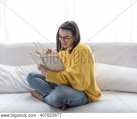 Stylish Brunette Girl In A Yellow Sweater And Glasses Reads A Book At Home On The Couch. The Concept