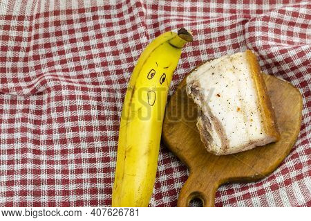Banana With A Face And A Piece Of Salty Fat On A White And Red Checkered Cloth. Ripe Banana With A G