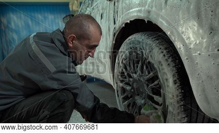 A Caucasian Man Works In A Car Wash Service. A Man In A Workers Uniform Washes The Wheel Of A Car Th