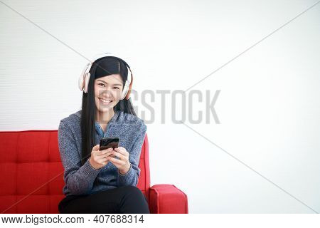 A Beautiful Smiling Young Woman Holding A Smartphone And Wearing Headphones To Music Sit On The Sofa