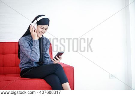 A Beautiful Smiling Young Woman Holding A Smartphone And Wearing Headphones To Music Sit On The Sofa