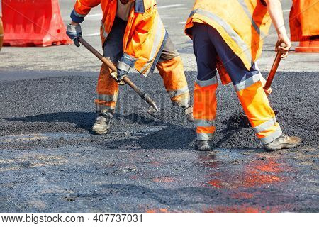 A Team Of Workers In Orange Overalls Use Shovels To Level The Asphalt Layer To Repair A Section Of T