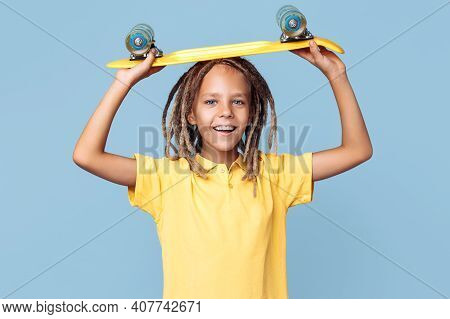 Positive Little Boy With African Dreads Holding Skateboard On The Head Over Blue Background.