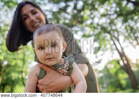 Young Brown-haired Mother Shakes Her Baby In Her Arms In The Park Among The Trees, Green Grass. Woma