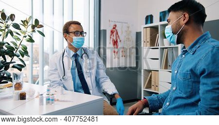 Young African American Handsome Male Physician Specialist In Medical Mask Talking With Man Patient E