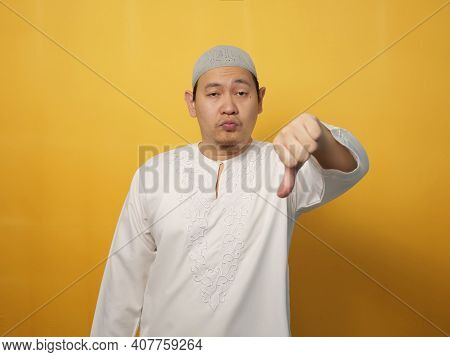 Portrait Young Asian Muslim Man Shows Upset Expression With Thumbs Down, Against Yellow Background