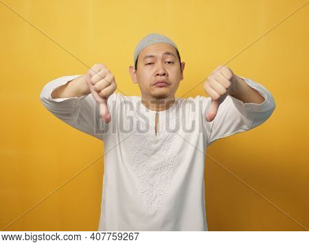 Portrait Young Asian Muslim Man Shows Upset Expression With Thumbs Down, Against Yellow Background