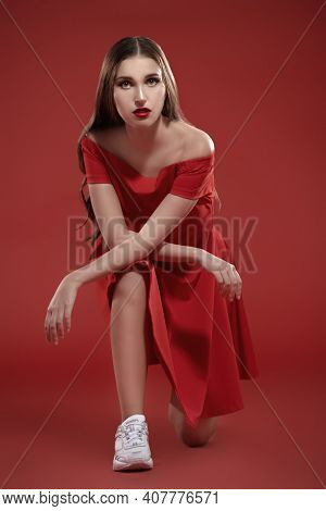 Beautiful brunette girl posing at studio in red dress and white sneakers. Red background. Sport chic style.