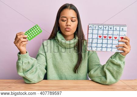 Beautiful hispanic woman holding birth control pills making fish face with mouth and squinting eyes, crazy and comical. 
