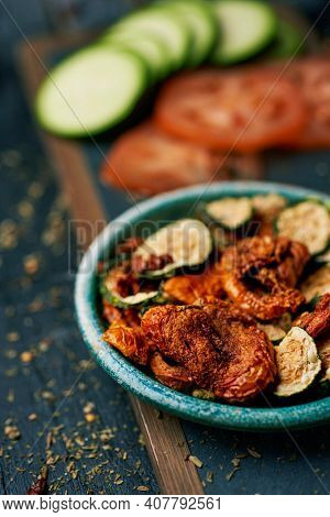 closeup of some slices of dried tomato and zucchini, served as appetizer or snack in a rustic green plate, and some slices of raw tomato and zucchini, on a dark gray rustic wooden surface