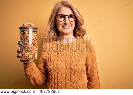 Middle age beautiful blonde woman holding jar of Italian dry pasta over yellow background with a happy face standing and smiling with a confident smile showing teeth