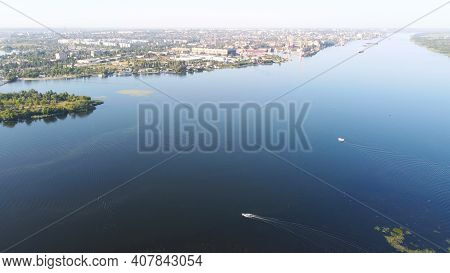 Drone Fly Over Waving River Of Blue Color Surrounded By Local Village With Various Buildings And Wet