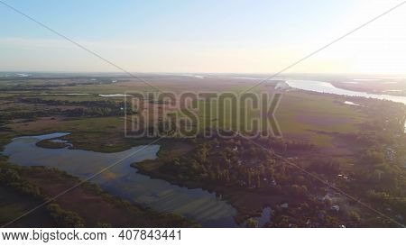 Drone Fly Over Waving River Of Blue Color Surrounded By Local Village With Various Buildings And Wet