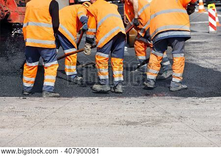 A Team Of Road Workers In Orange Reflective Uniforms Use Shovels To Level Fresh Asphalt On A Clear D