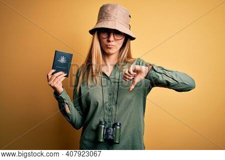 Blonde tourist woman with blue eyes on vacation holding australian passport using binoculars with angry face, negative sign showing dislike with thumbs down, rejection concept