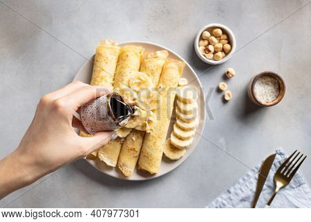 Womans Hand Pours Date Syrup Over Thin Banana Pancakes. Healthy Vegan Breakfast. Gray Background. To
