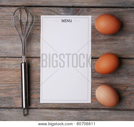 Menu sheet of paper with egg whisk and eggs on rustic wooden surface background