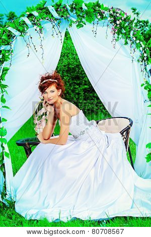 Beautiful bride with chaming red hair sitting under the wedding arch. Wedding dress and accessories. Wedding decoration. 