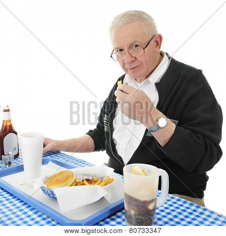 A senior adult man looking up at the viewer as he enjoys a french fry from his fast food basket.  On a white background.