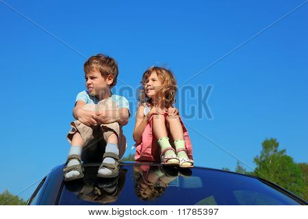 Liten pojke och flicka sitter på biltaket på blå himmel, händerna på ben