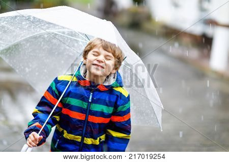 Beautiful little kid boy on way to school walking during sleet, rain and snow with an umbrella on cold day. Happy and joyful child in colorful fashion casual clothes