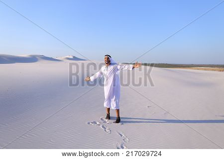 Stately young sheikh walks on foot along bottomless desert leaving traces on sand, enjoying warmth of day. Attractive Muslim smiles widely and spreads hands to sides, straightens clothes in wind and whirls around, delighted by breadth of desert. Swarthy, 