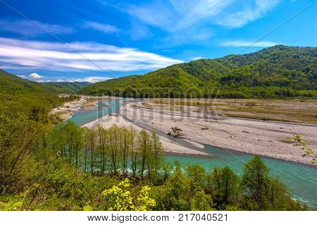 The azure mountain river flows around the stones between the forest and the mountains against the background of a bright blue sky in spring or summer. Shahe river, Bolshoy Kimchay, Sochi, Russia.