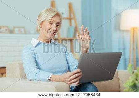 Progressive pensioner. Clever enthusiastic progressive senior woman waving her hand and feeling happy while using a video call in her laptop and talking with grandchildren