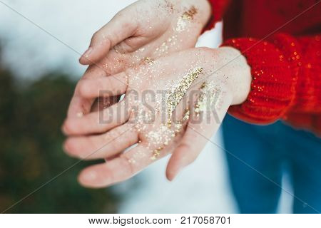 Young curly girl in crystal sunglasses blowing on hands with golden glitters on blue background.