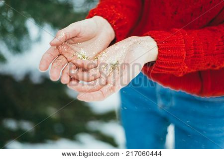 Young curly girl in crystal sunglasses blowing on hands with golden glitters on blue background.